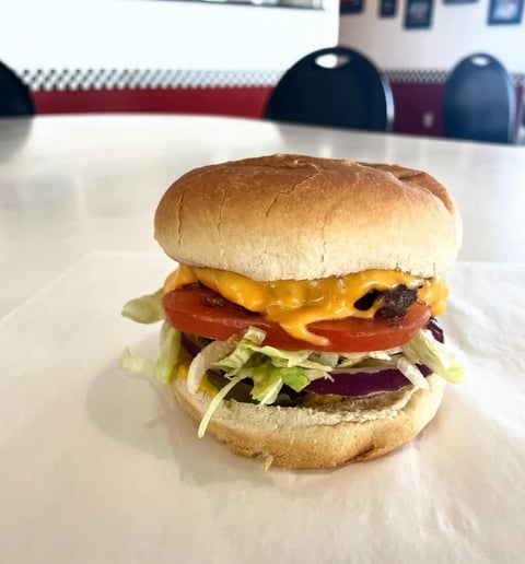 Gourmet burger with melted cheese, tomato, lettuce, and charred patty on a toasted bun, served on white paper in a restaurant setting