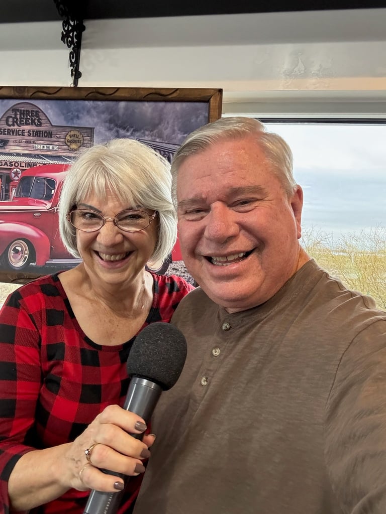 Two smiling people holding a microphone together in front of a vintage gas station sign backdrop