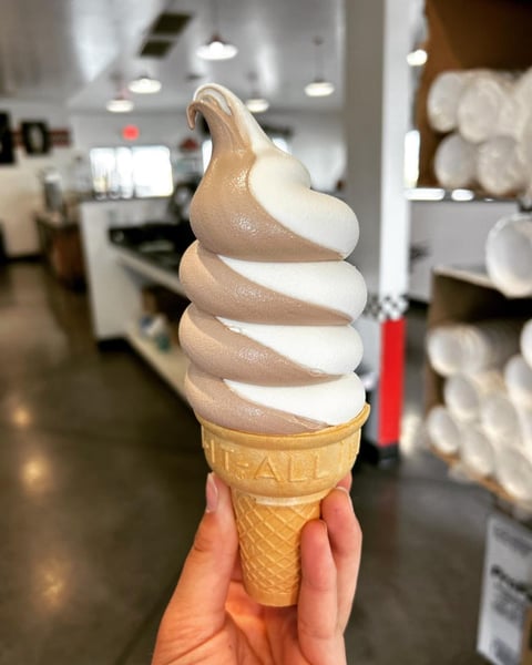 Hand holding a soft-serve ice cream cone with vanilla and chocolate swirl, in an ice cream shop